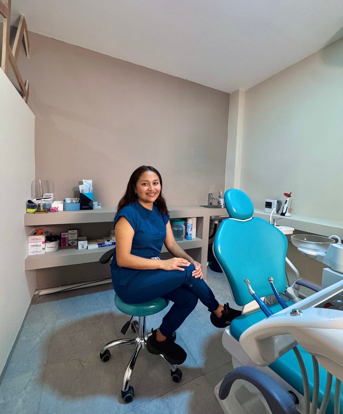 Female dentist in dark blue scrubs sitting on a stool in a dental examination room with sink and equipment visible