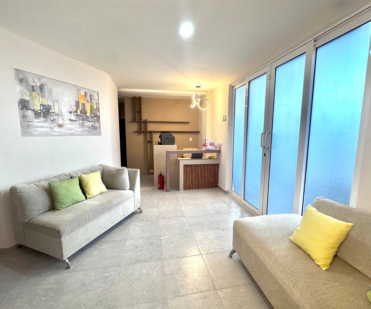 Modern apartment living room with two beige sofas, yellow accent pillows, large blue glass doors, kitchenette, and framed artwork on white walls